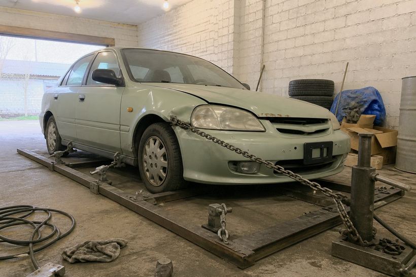 Старая светло-зелёная Honda Civic закреплена захватами на рамном стапеле, от угла рамы к переднему крылу натянута стальная цепь